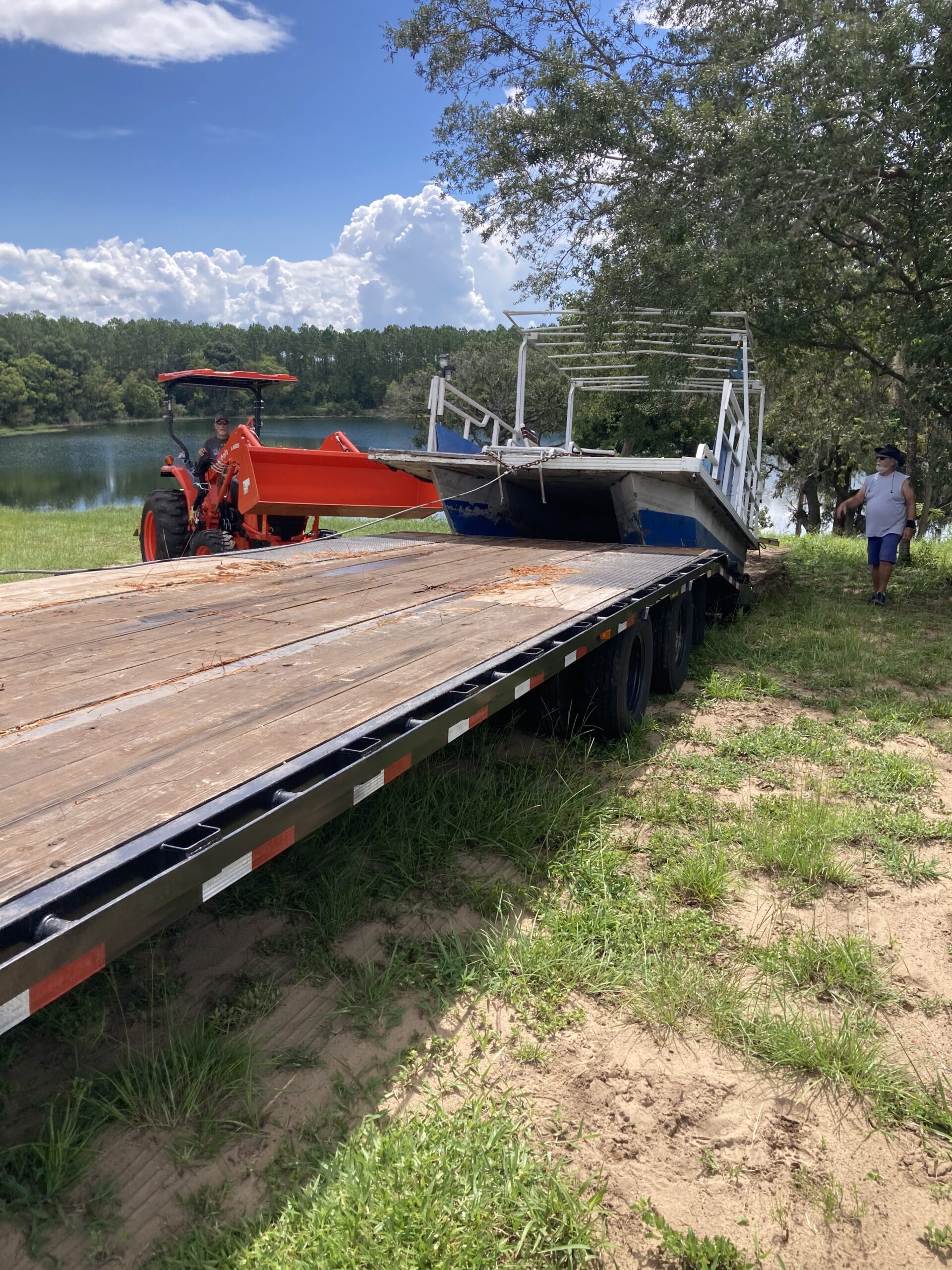Tractor loading a large boat onto a flatbed trailer during a complete property cleanout near a lake